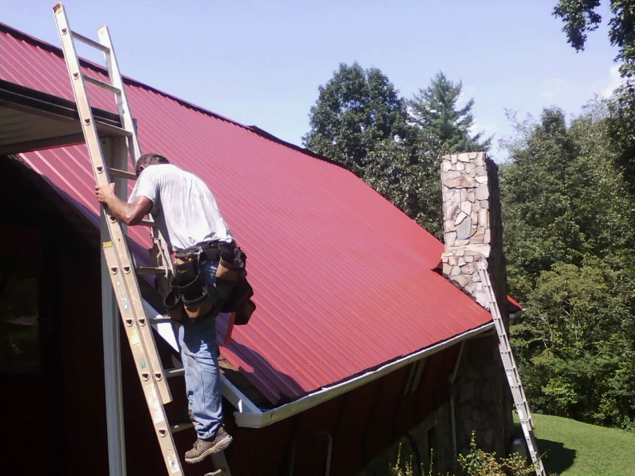 Roofer on a ladder beside a red metal roof and stone chimney — Soffit & Fascia Repair by Roofing Brothers in Pickerington
